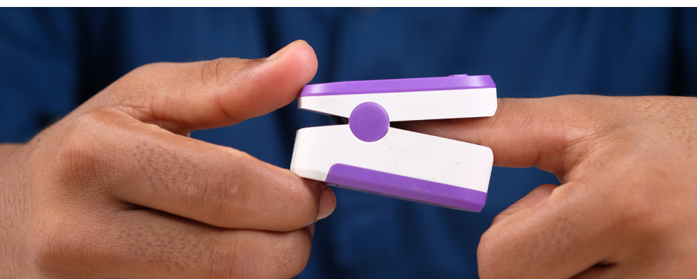 A digital pulse oximeter displaying oxygen saturation and pulse rate, placed on a wooden surface beside a stethoscope in a clinical setting in Pakistan.